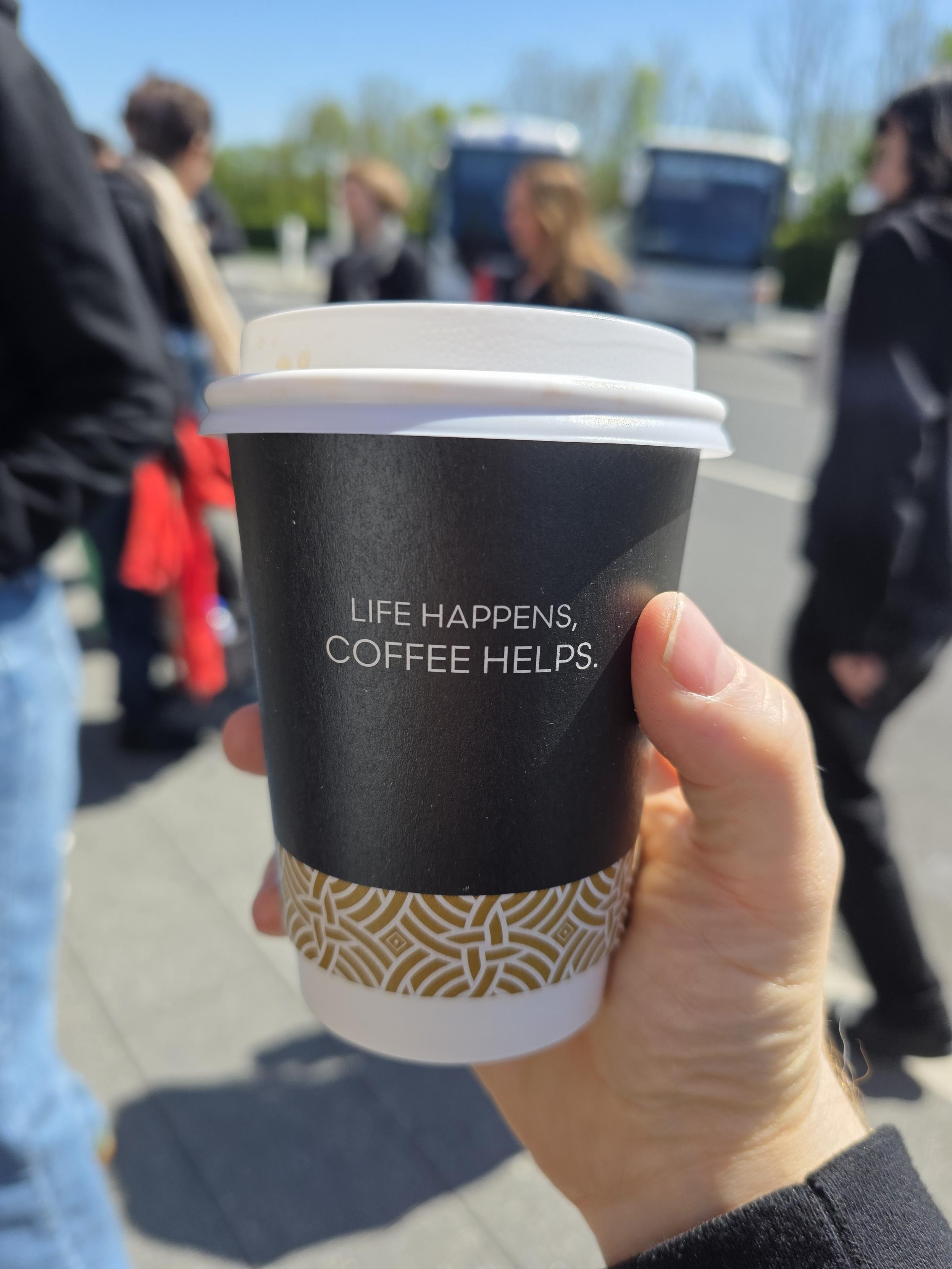 A black coffee cup with a white lid, against a backdrop of people.

The Coffee cup reads "life happens, coffee helps"